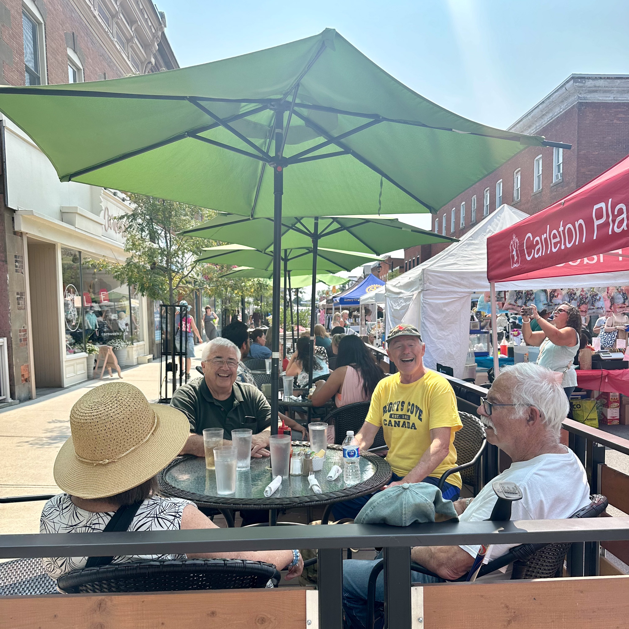 A group smiling under a patio umbrella while the festival continues in the background.