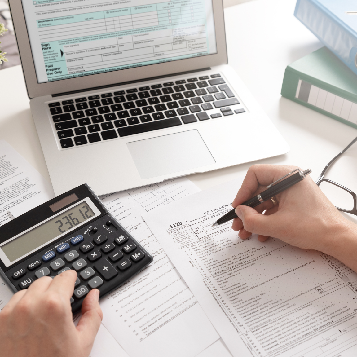 Calculator displays numbers, a hand holds a pen filling out forms and a laptop is open on the desk.